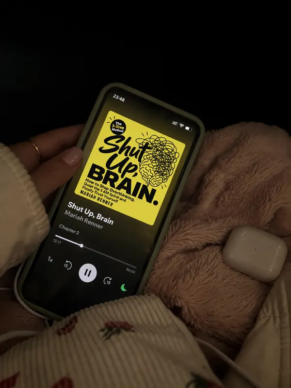 At night in bed, a hand holds a phone that plays chapter 2 of Shut Up, Brain with a sleep timer set, an earbuds case on the blanket.