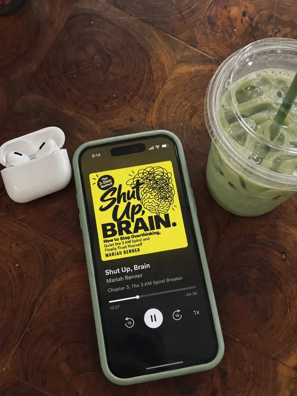 A phone in a sage green case on a wooden table plays the Shut Up, Brain audiobook, next to an iced matcha and an open earbuds case.