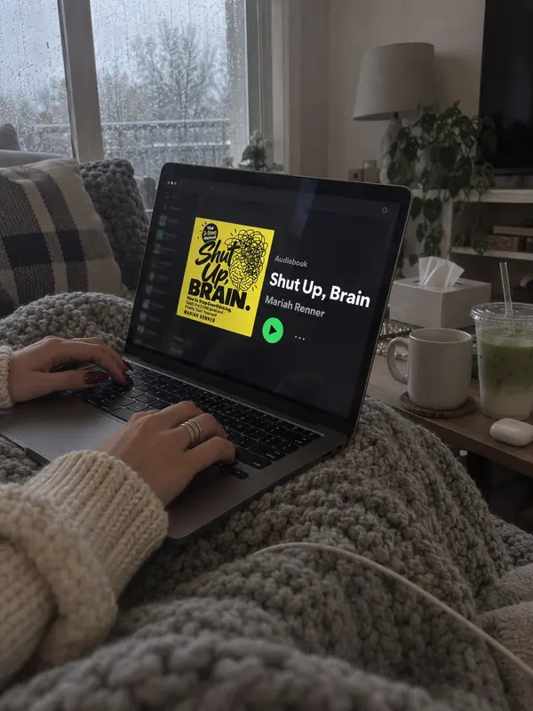 A laptop on a knitted blanket by a rainy window shows the Shut Up, Brain audiobook page, an iced matcha on the side table.