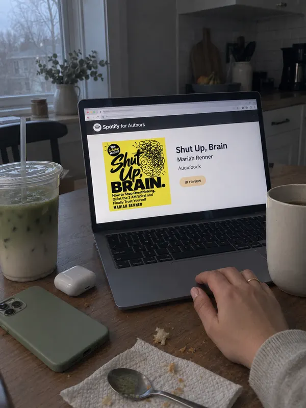 A laptop on a kitchen table showing the Shut Up, Brain audiobook marked in review, next to an iced matcha, a mug of coffee, an earbuds case and a phone lying face down. One hand rests on the trackpad.